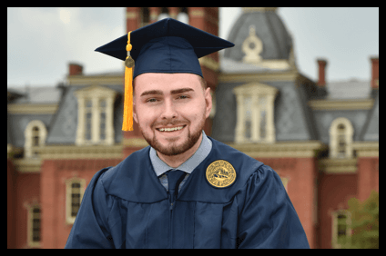 Male college graduate in a cap and gown smiling in front of West Virginia University’s campus.