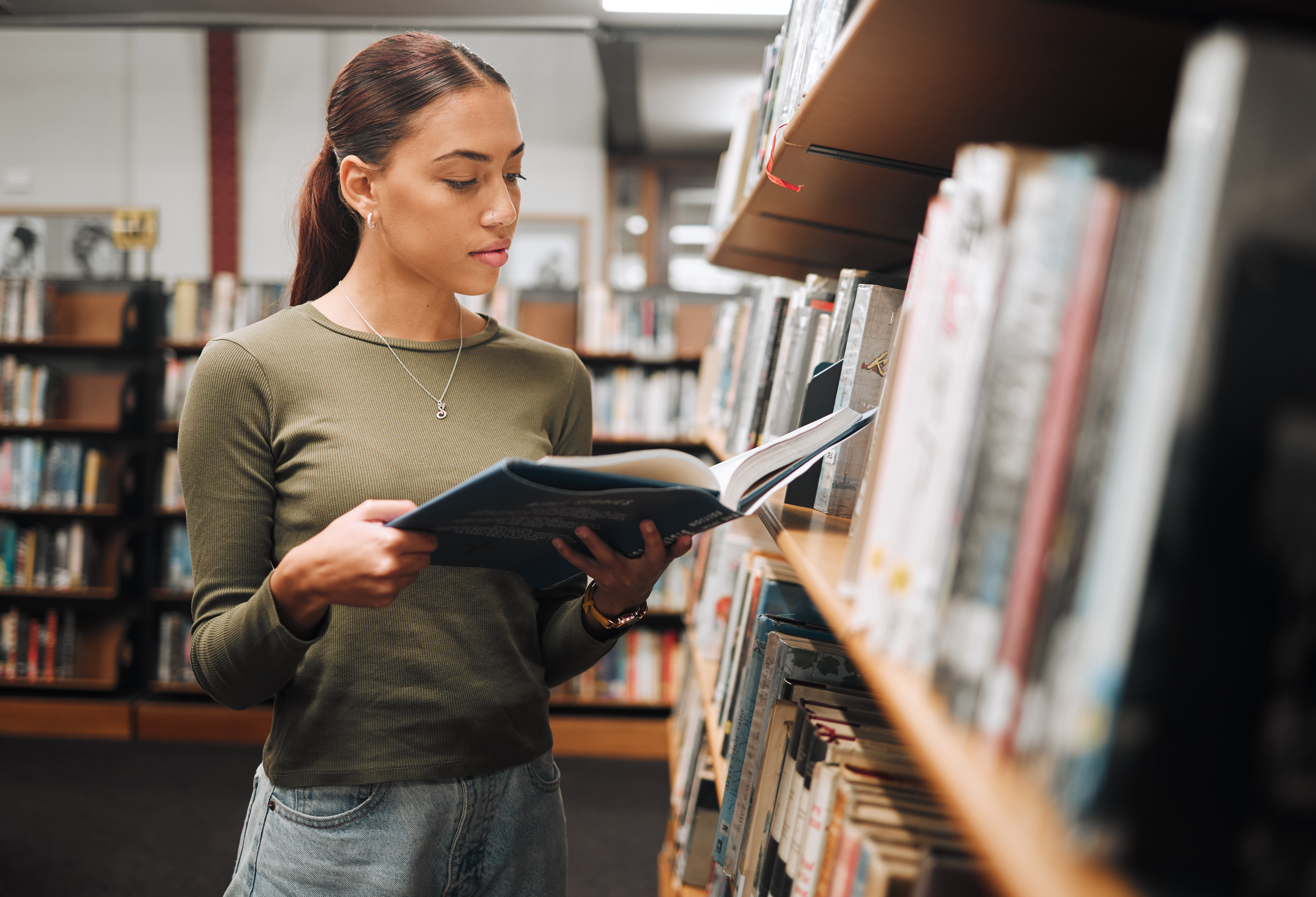 A woman in a green top reads a book in a library aisle, surrounded by shelves filled with books.