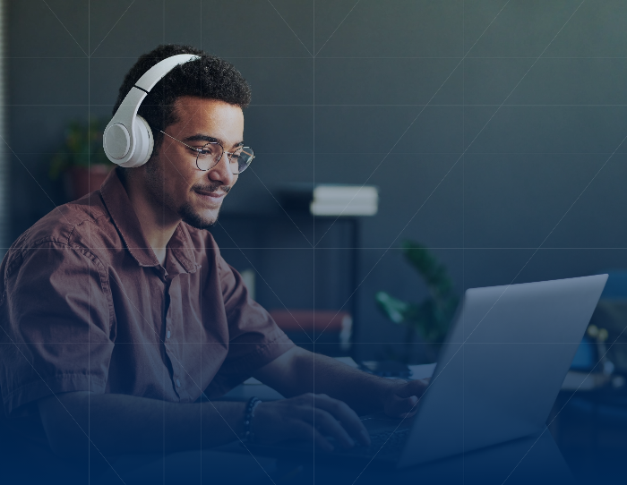 A young man wearing headphones and glasses focuses intently on his laptop screen in a modern office environment.