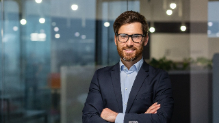 A bearded man with glasses wearing a dark suit stands with his arms crossed, smiling in an office setting with glass walls.