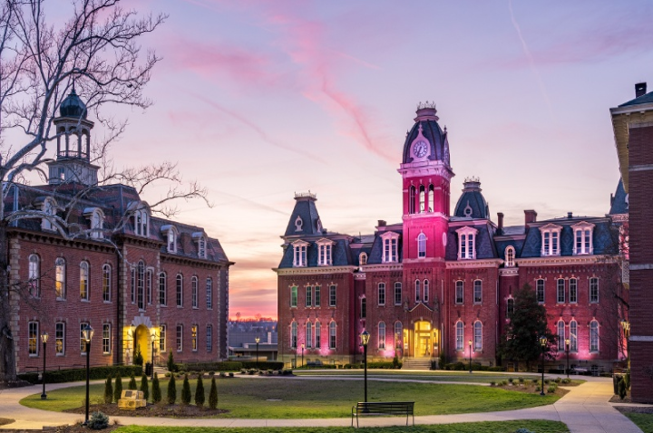 Campus buildings with historic architecture and a central tower are lit up at sunset, with a pink and purple sky in the background.