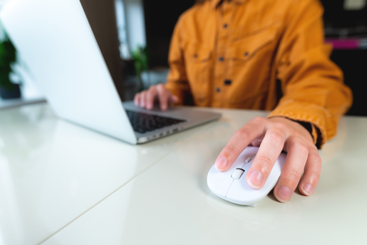 Person in an orange jacket using a laptop on a white desk, holding a computer mouse.