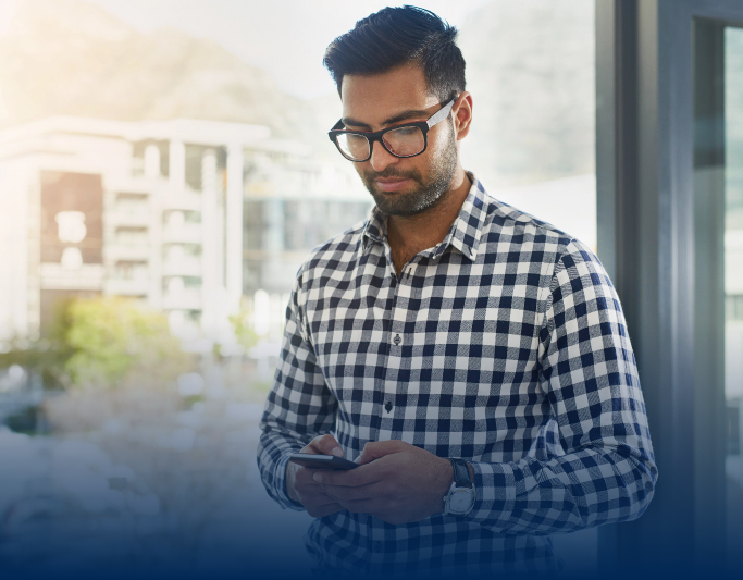 A man with glasses and a checkered shirt looks at his phone while standing indoors near a window.