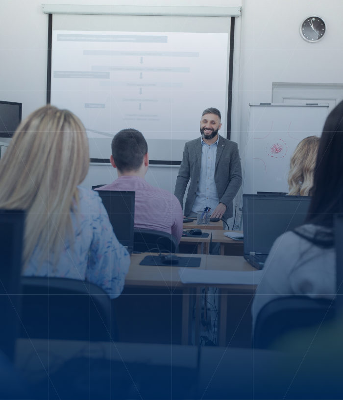 A male teacher standing and smiling at a classroom full of students with laptops, presenting a lesson on a whiteboard in the background.