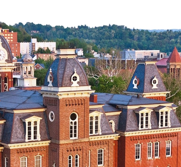 A historic brick building with multiple towers and dormer windows sits in the foreground, with a green, tree-covered hillside and additional buildings visible in the background.